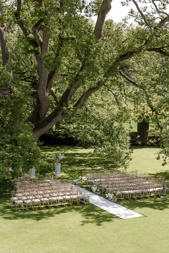 Chairs set out on the grass under a great spreading tree, a white aisle down the middle