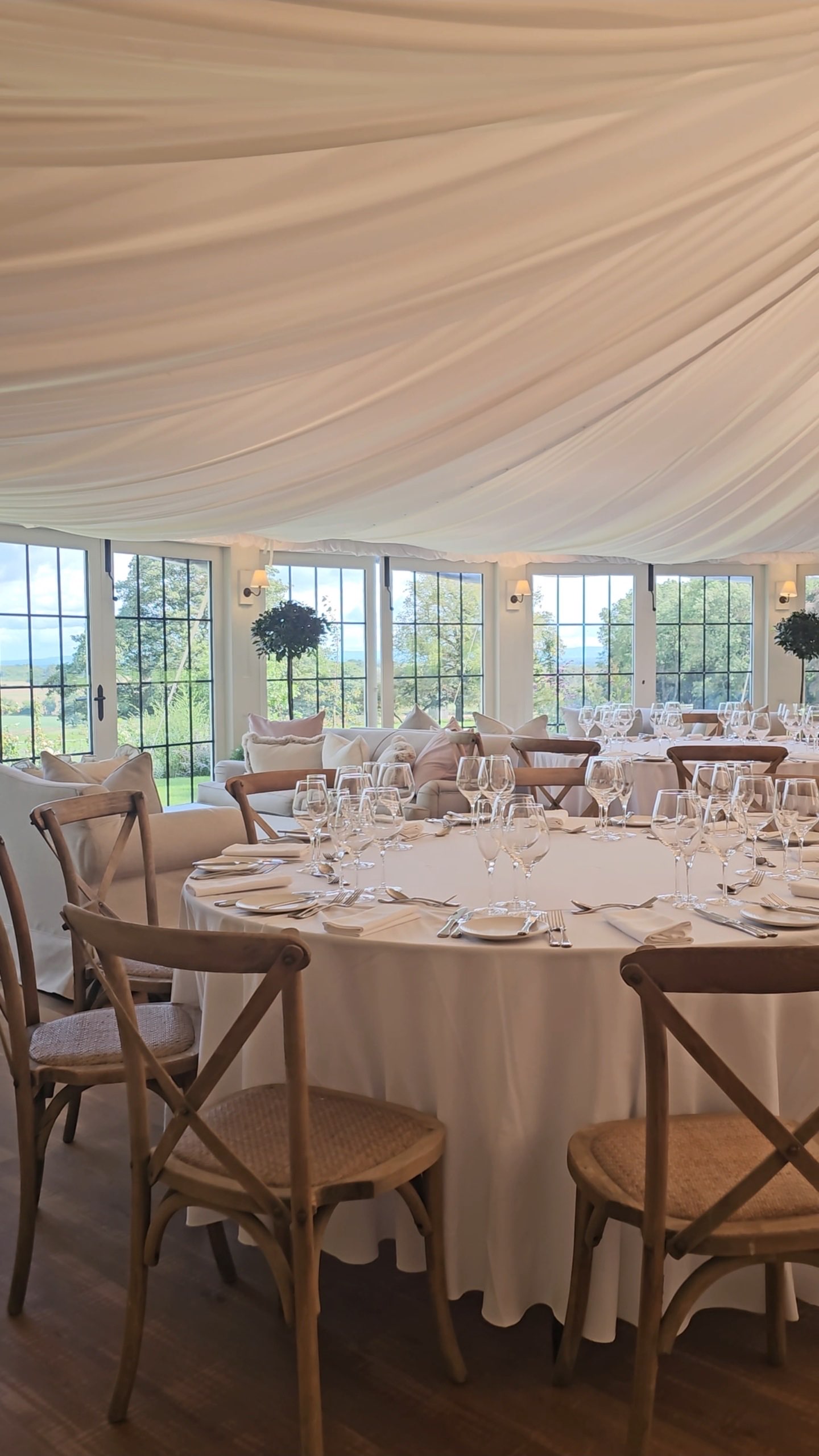 Inside the marquee: a round table laid with glasses under a draped ceiling, tall windows onto the fields
